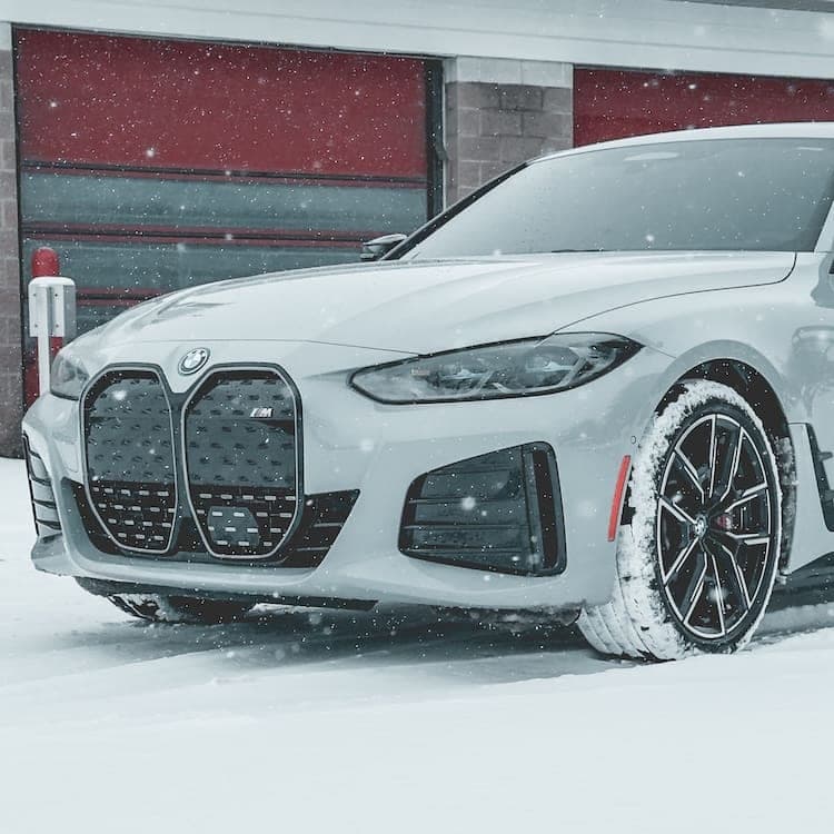 Front three-quarter view of a light grey BMW i4 M50 in falling snow, red garage doors behind it.