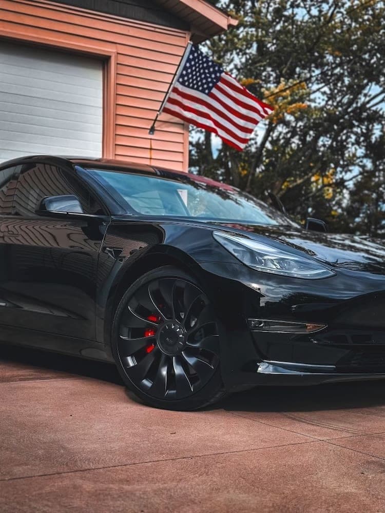 A black Model 3 with black wheels and red calipers parked under an American flag.
