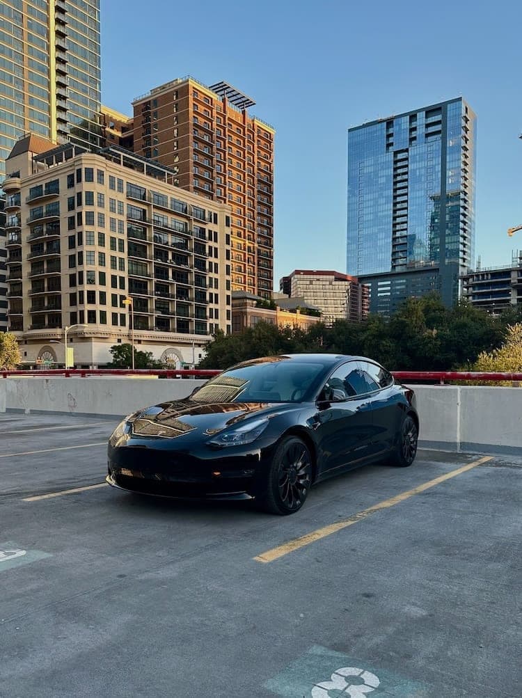 The black Model 3 on a rooftop parking deck with city towers and blue sky behind.
