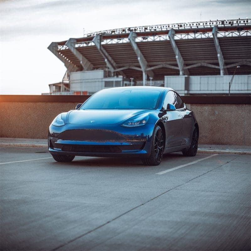 The blue Model 3 in an empty parking lot with a stadium in the background.