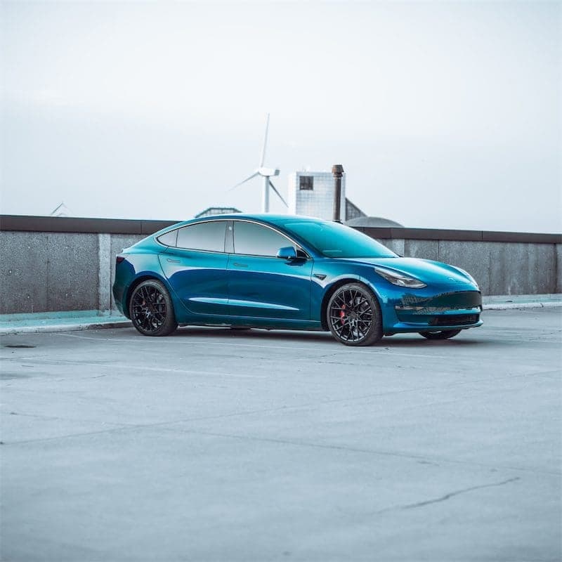 Side view of the blue Model 3 on a rooftop deck, a wind turbine in the distance.
