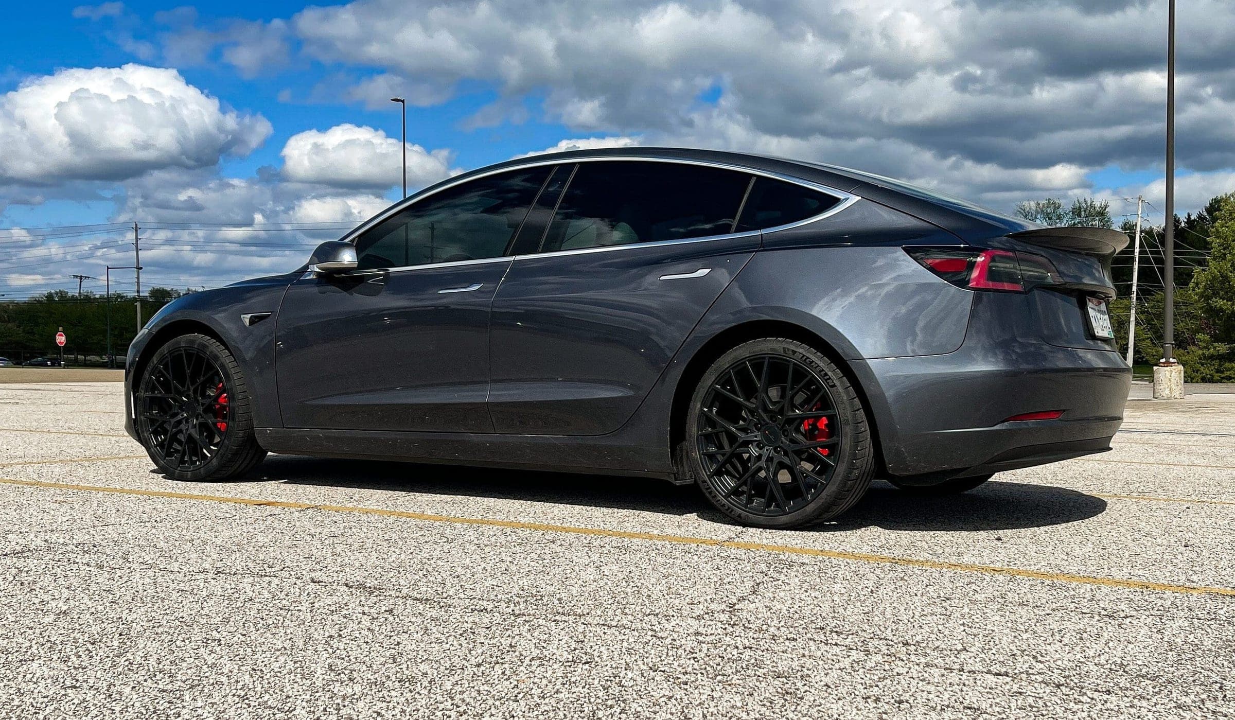 Side view of a grey Model 3 on black wheels in a parking lot under a cloudy sky.