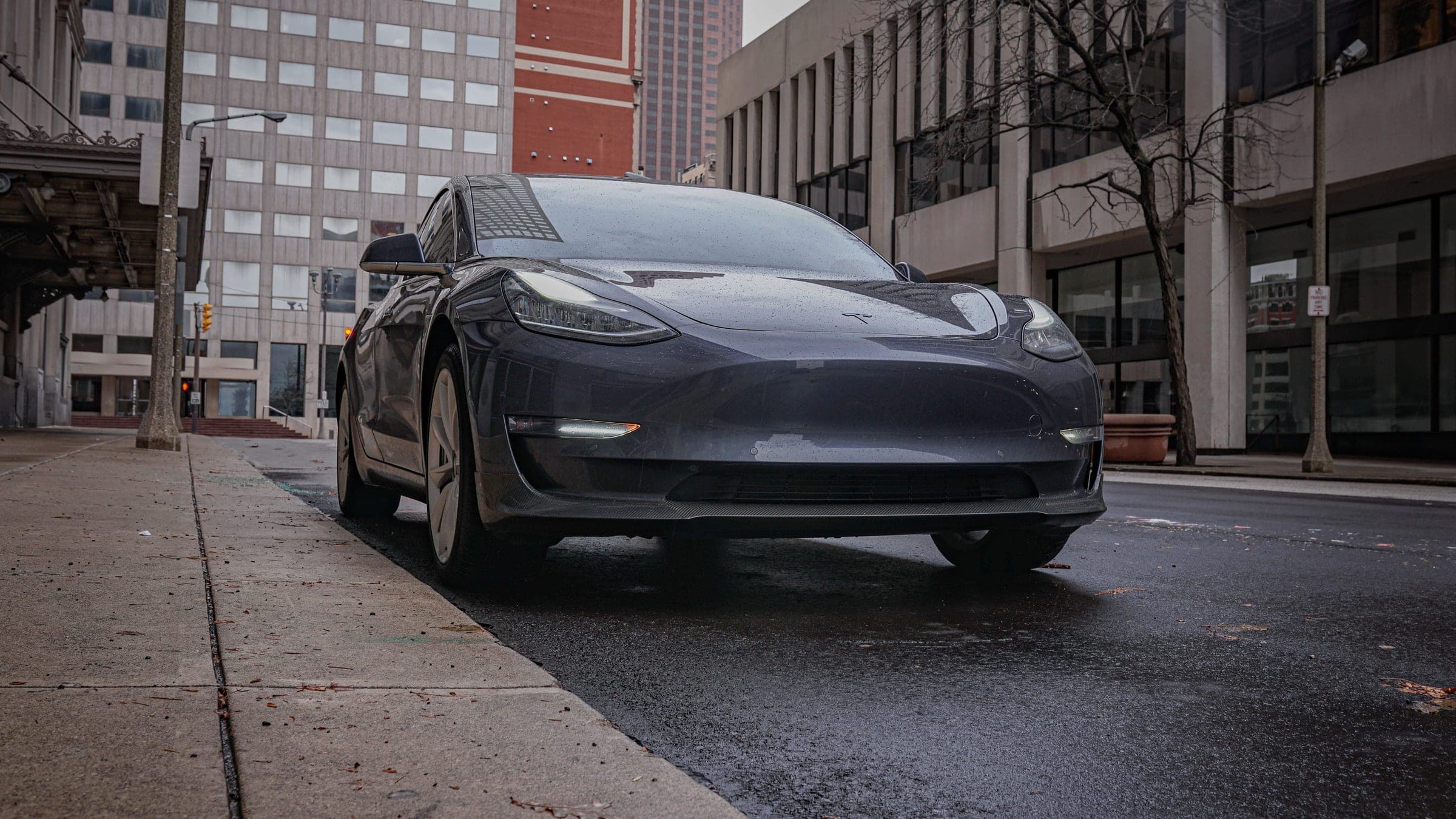 A grey Model 3 parked at the curb on a downtown street, low angle.